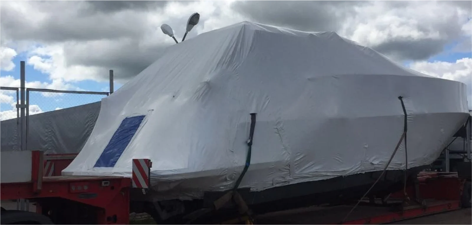 Liela motorjahta, droši iepakota baltā aizsargplēvē, uzkrauta uz sarkanas zemas platformas piekabes negabarīta kravu pārvadāšanai no Itālijas uz Norvēģiju, ko veic Lazarus.